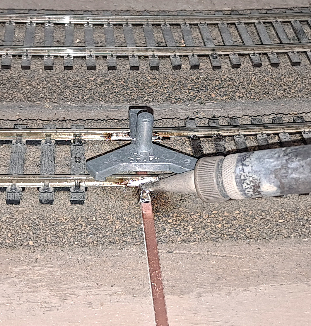 Three-point rail gauge above joint in track with PCB tie under joint and pencil-type soldering iron tip coming in from right.