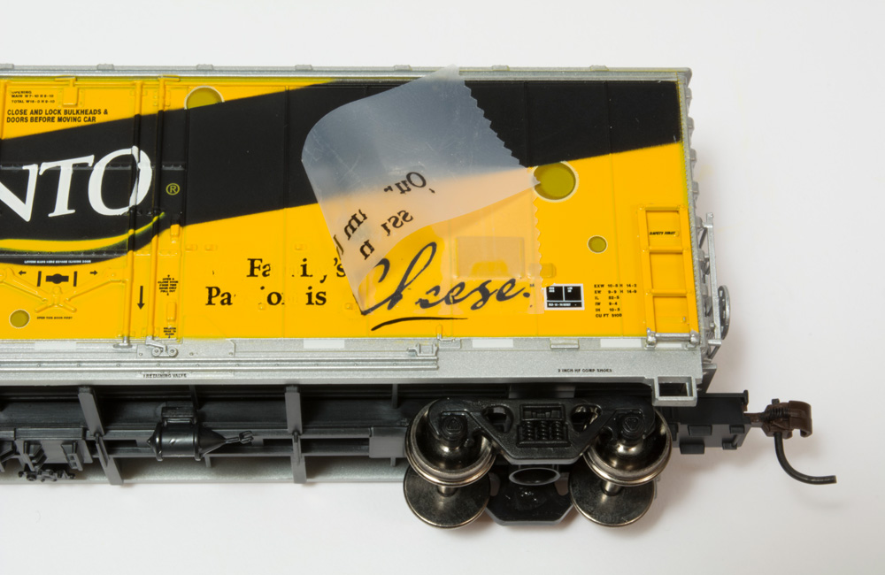 Scotch tape is partially peeled back from the side of a yellow-and-black boxcar model, lifting some of its black printing.