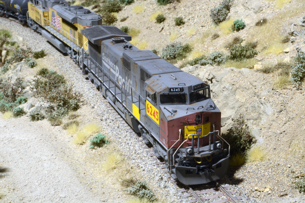 Photo of gray and red HO scale locomotive with yellow patches on a scenicked model railroad. 