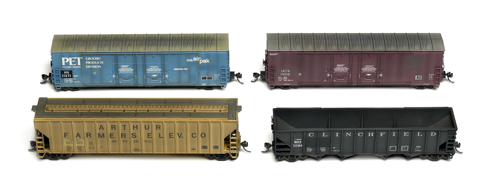 Photo of HO scale yellow covered hopper, black quad hopper, blue boxcar, and maroon boxcar with patchouts on white background.