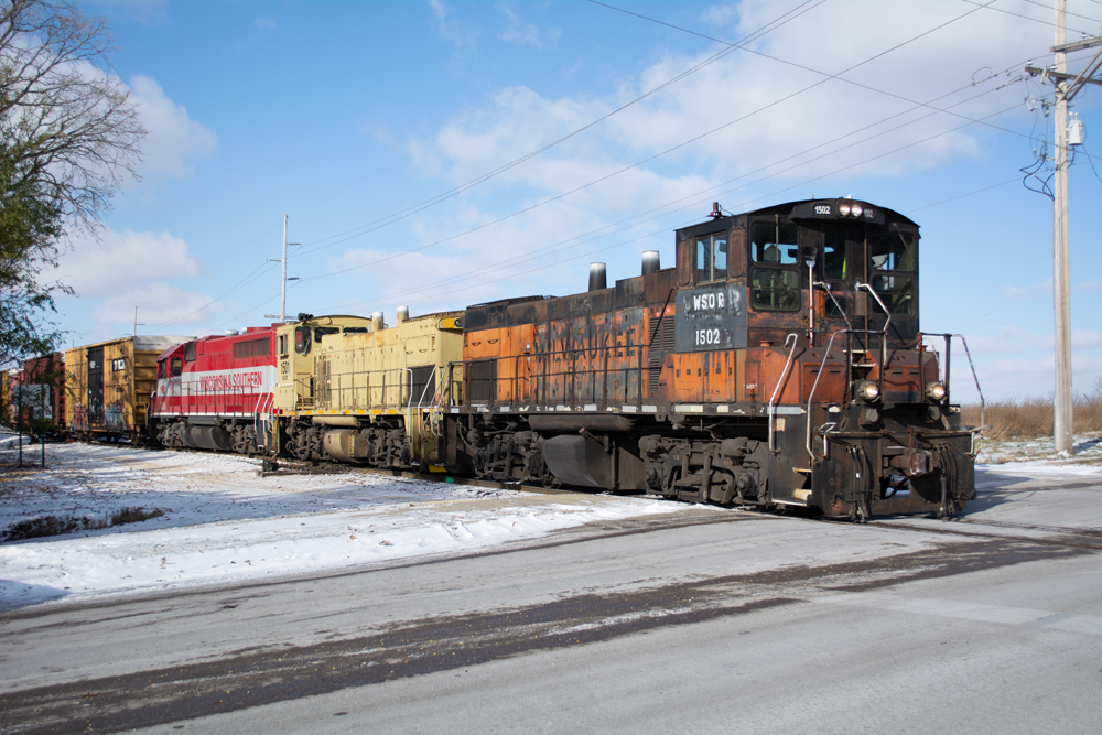 Photo of orange-and-black, yellow, and red-and-gray locomotives switching cars on a partly cloudy winter day with snow on the ground.