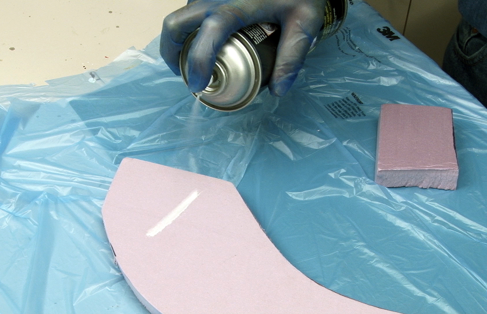 A rubber-gloved hand holds a can of spray adhesives while applying the adhesive to a pink foam board carving