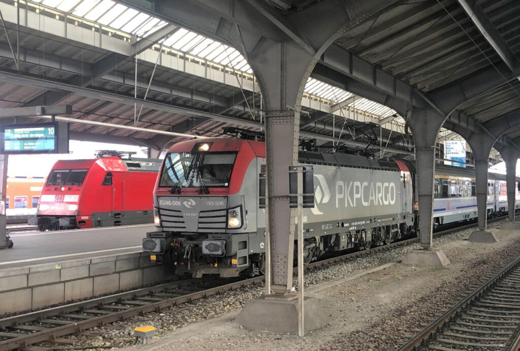 European passenger trains, one with red locomotive, one with gray locomotive, sit at station platform