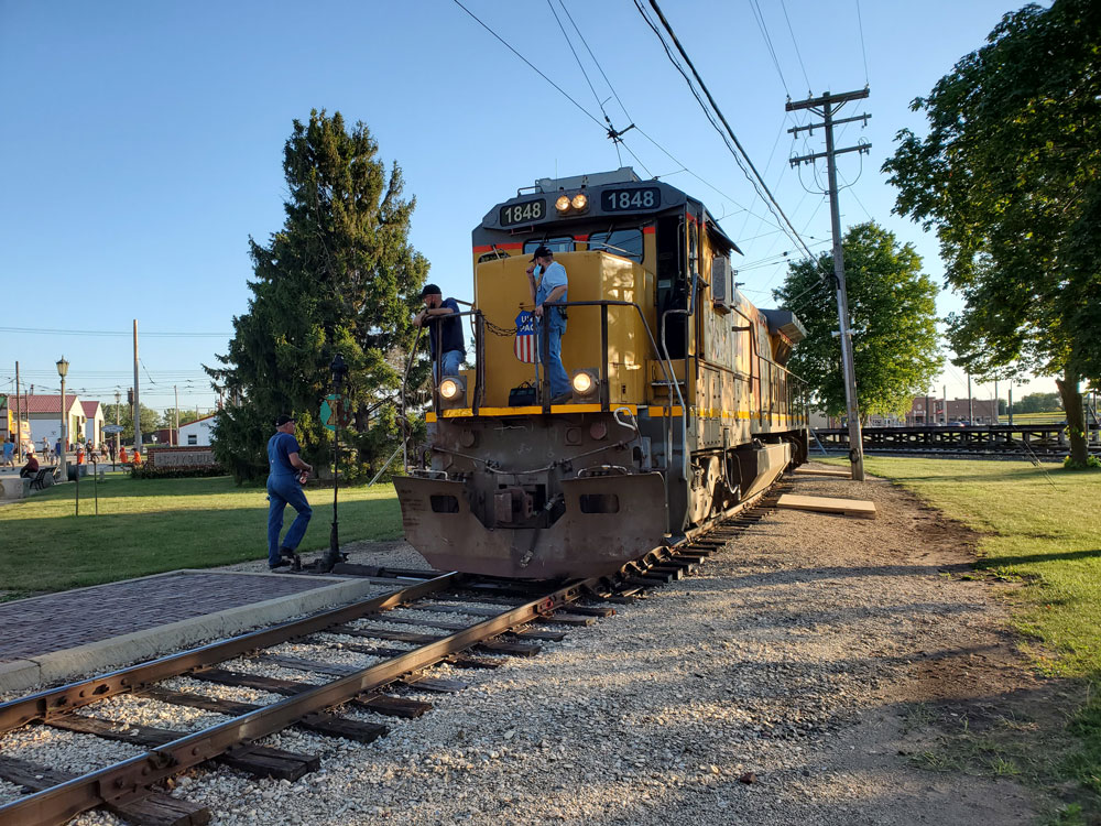 Yellow locomotive on tracks with two people on the pilot and one person next to tracks