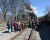 Large group of people stand near tracks and the front of a stopped steam engine to pose for picture