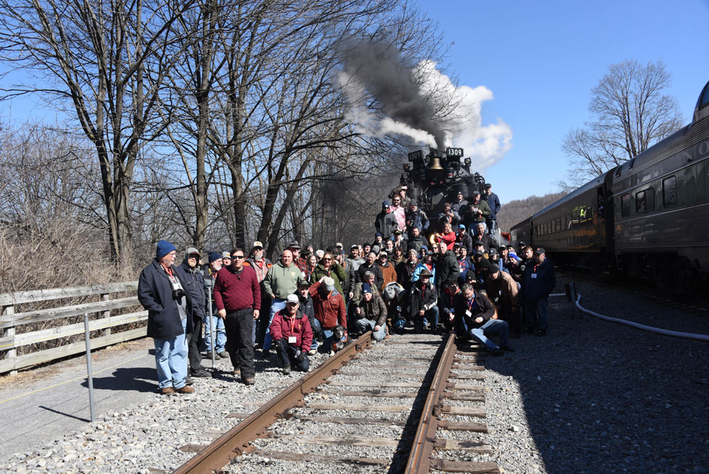 Large group of people stand near tracks and the front of a stopped steam engine to pose for picture