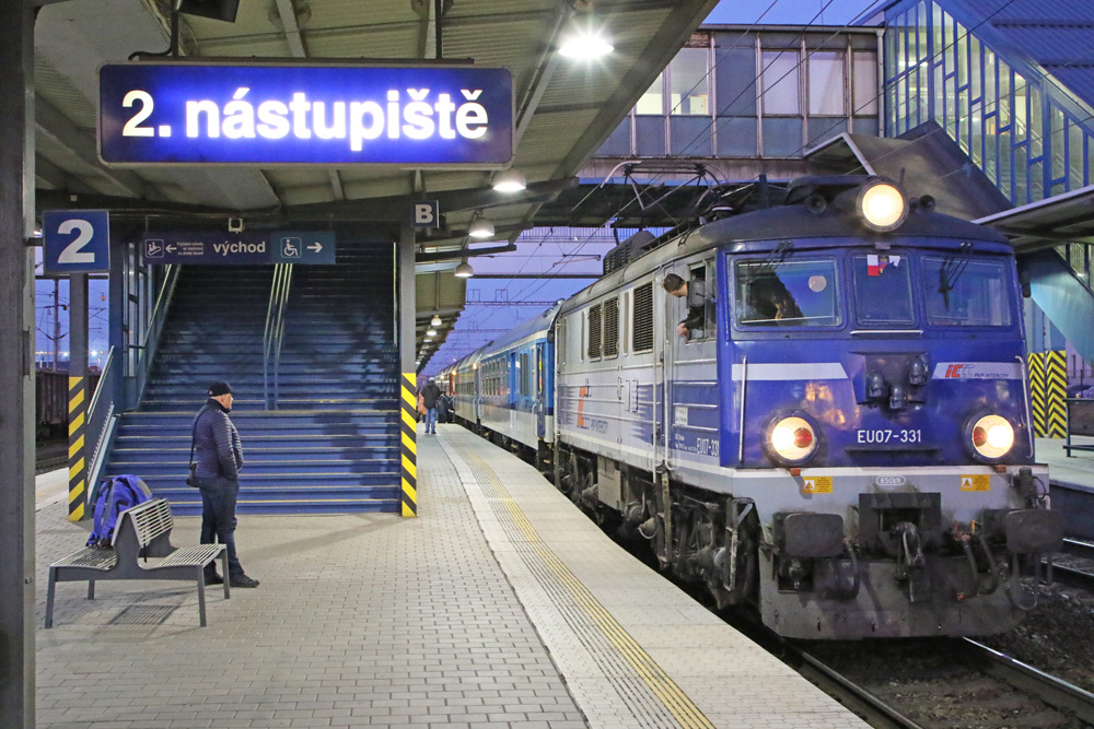 Train with blue and gray locomotive arrives at station with one person on platform