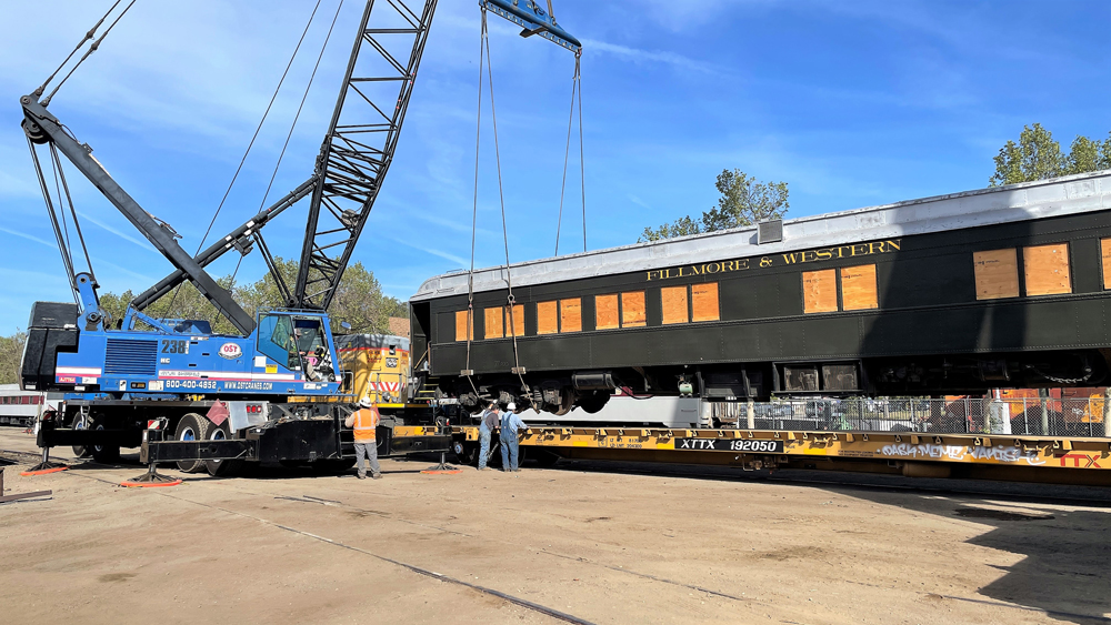 Green heavyweight passenger car being lifted by crane onto flatcar