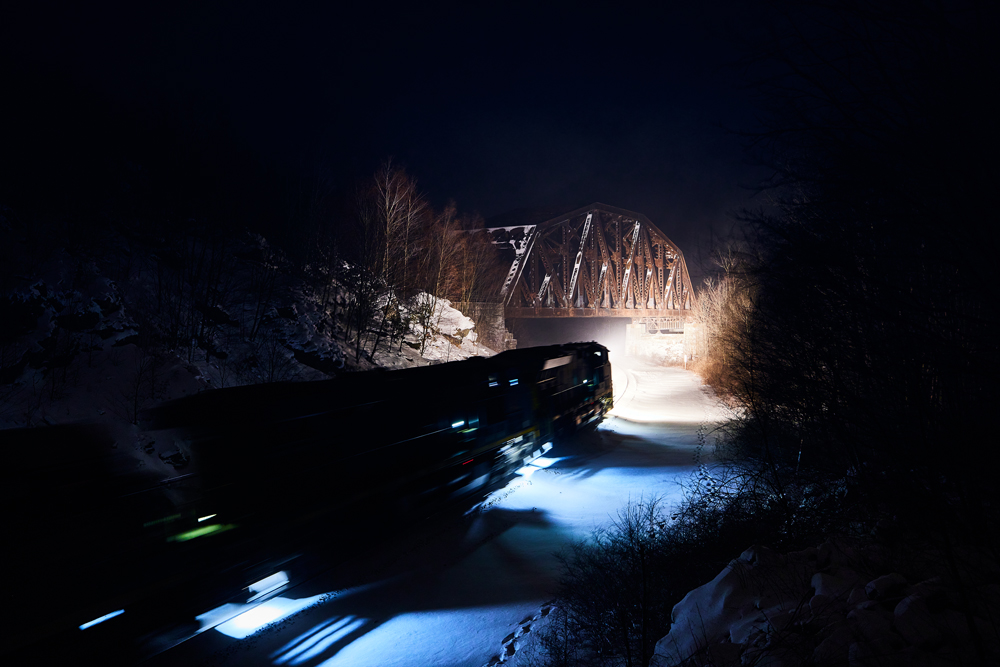 A train bridge illuminated by the headling of a train passing underneath at night, in a snowy scene.