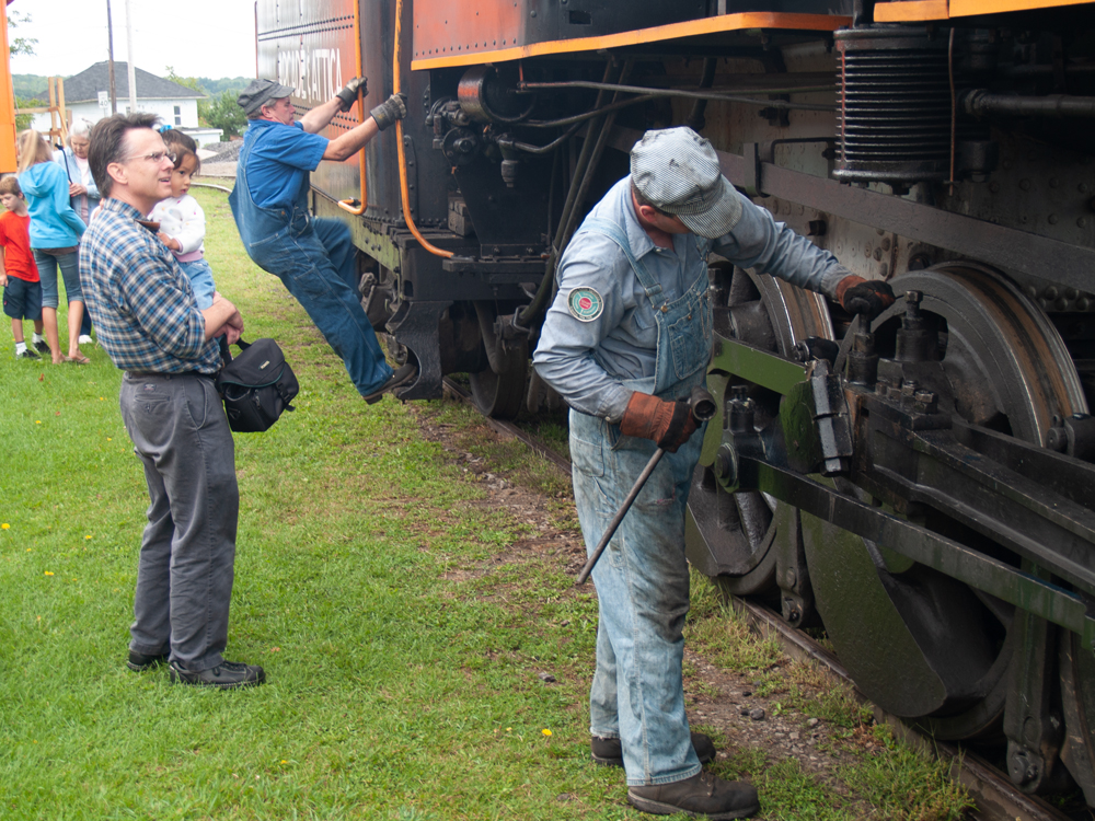 Man holding child watching crew member work on steam engine