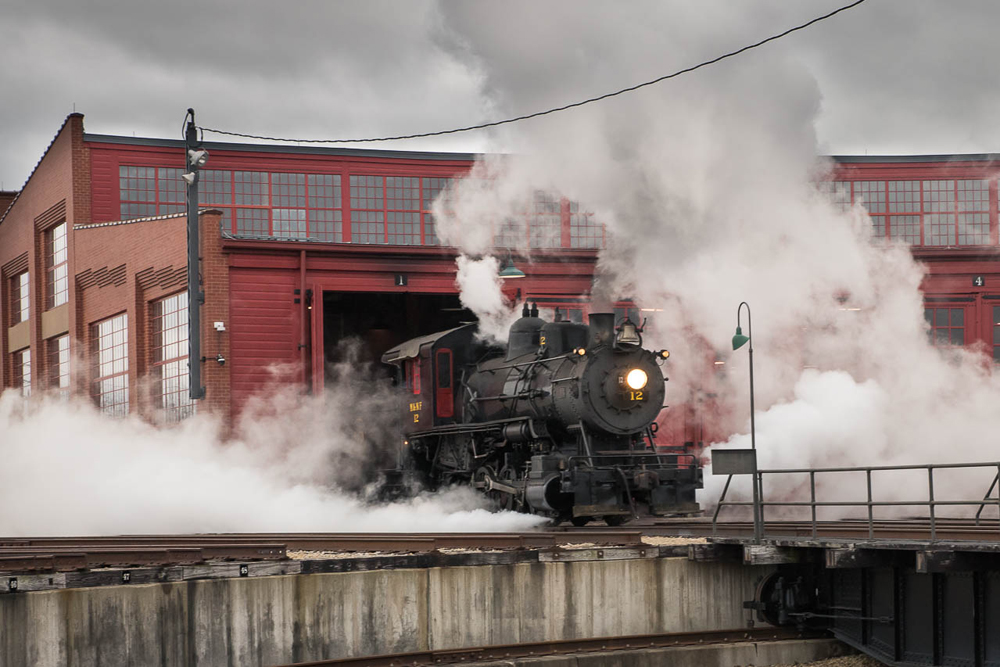 Small steam locomotive in clouds of steam outside roundhouse