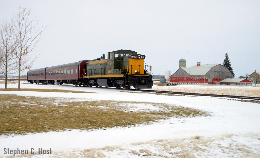 Diesel locomotive pulling two passenger cars passes barn