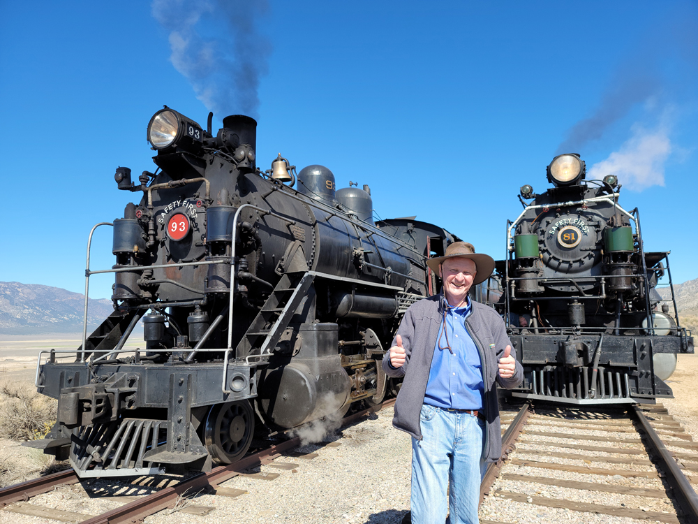 Smiling man giving thumbs-up signal while standing in front of two steam locomotives