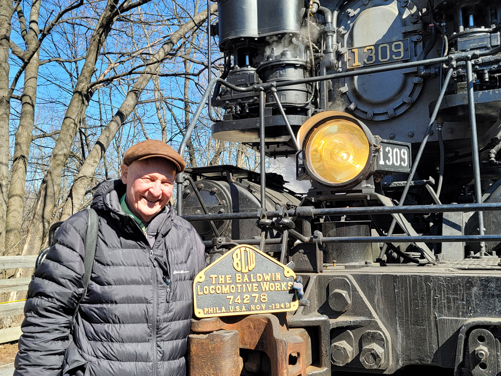 Man posing with builders' plate in front of steam locomotive