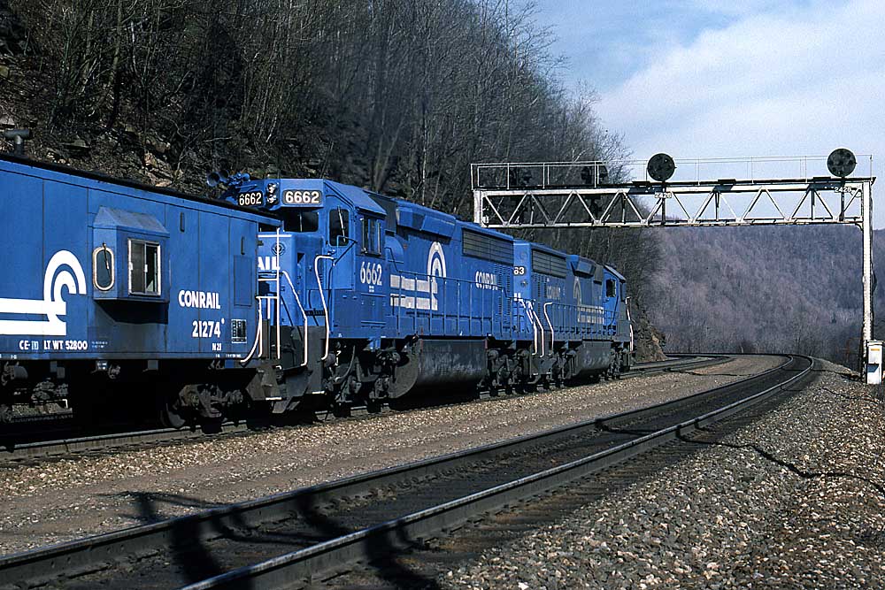 Two blue Conrail locomotives behind caboose under signal bridge