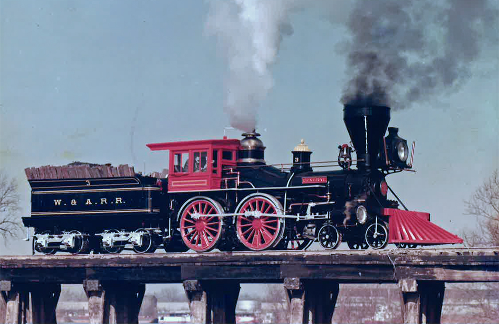 An ornate, old-fashioned black-and-red 4-4-0 steam locomotive smokes on a wood trestle