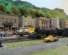 A large black steam locomotive waits while another pulls out of a yard in a mountain town