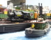 A gray and green steam tugboat moves toward a car float on which a small saddle-tank steam switcher smokes