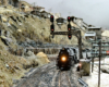 A steam locomotive with a coal train rolls under a signal bridge with a coal tipple in the distance