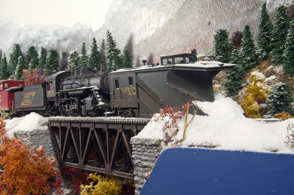 Snowplow attached to train moving snow over a bridge on a model railroad