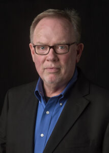 Man with dark suit coat and open collared blue shirt.