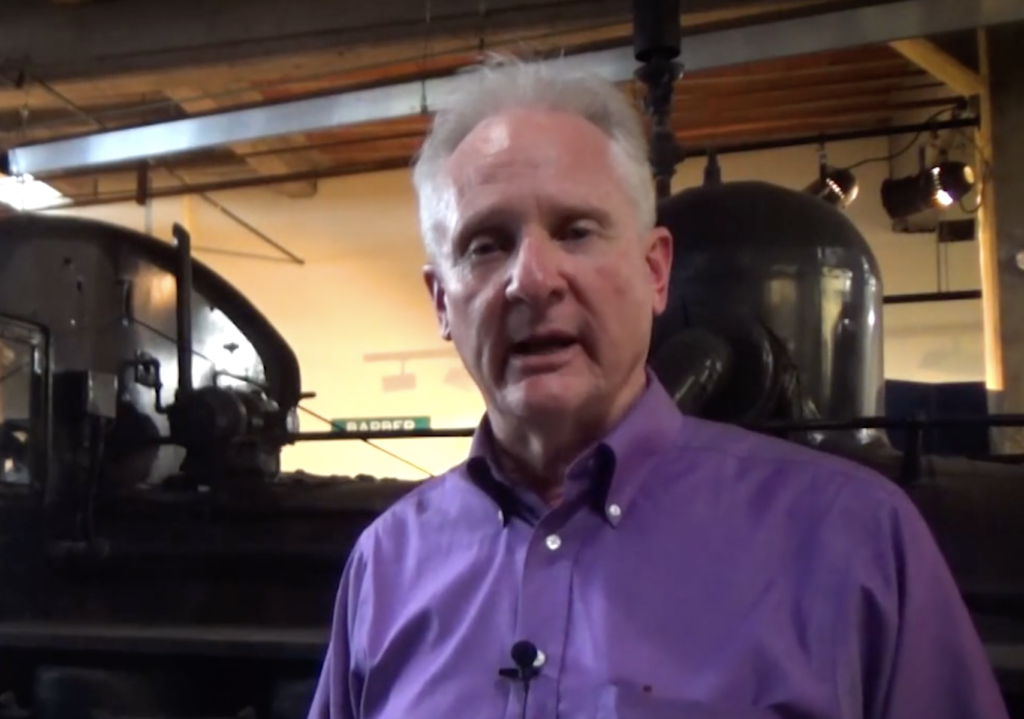 Man speaking in front of steam locomotive.