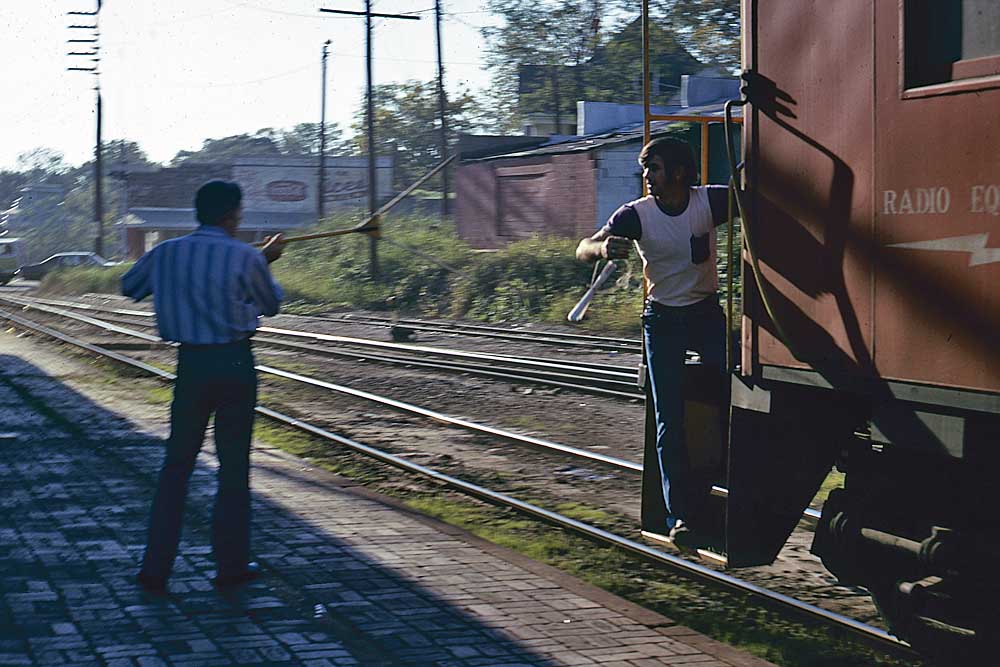 Man prepares to catch papers hung out by man on passing freight train caboose