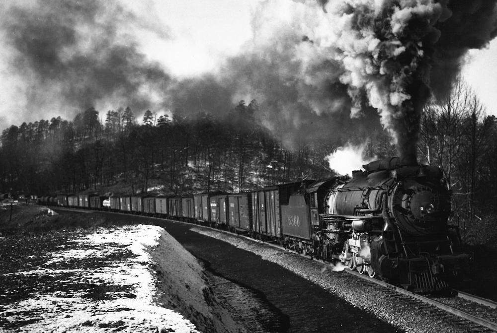 Heavy steam locomotive with freight train in the mountains surounded by trace amounts of snow.