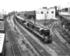 Two diesel locomotives lead a freight train through a rail yard and under a highway overpass.