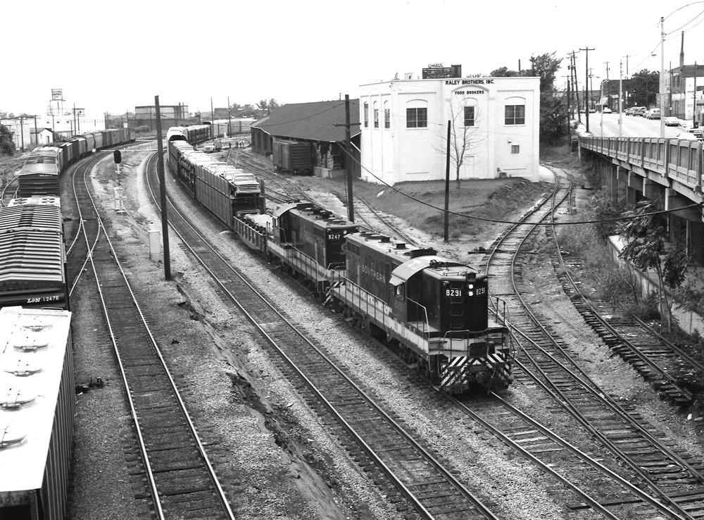 Two diesel locomotives lead a freight train through a rail yard and under a highway overpass.