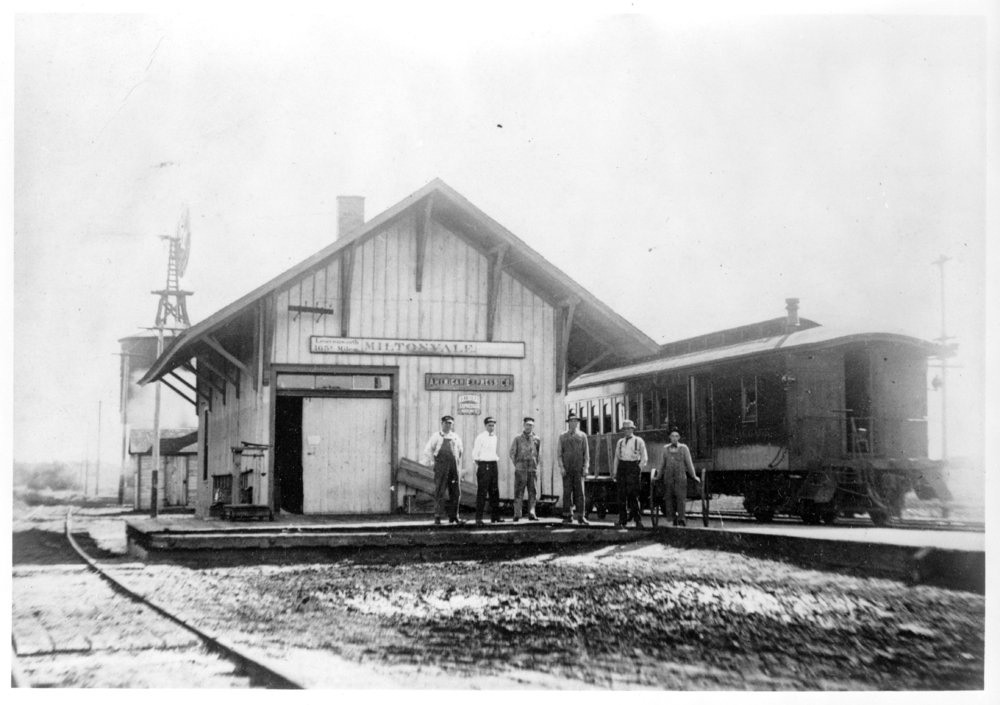 Black and white photo of a railroad depot, with six standing men and a passenger car nearby.