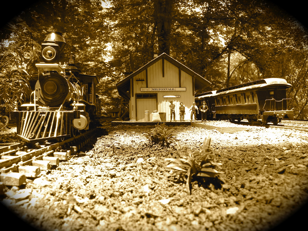 Garden railway scene with a steam locomotive, depot, and several figures in a sepia tone