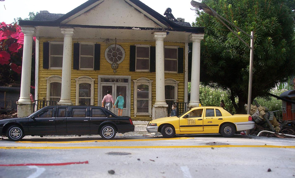 Model structure with two model cars in front of it, and two people walking nearby