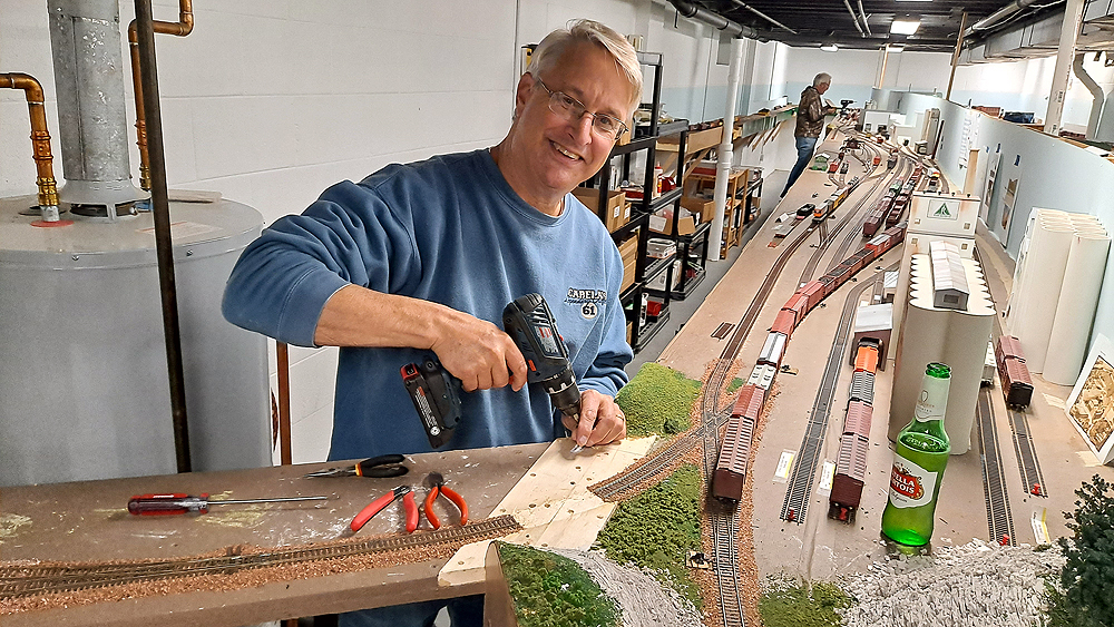 Packing up a model train layout: A man in a blue sweatshirt pauses while using an electric drill to remove screws from a duckunder connecting two parts of a layout together.