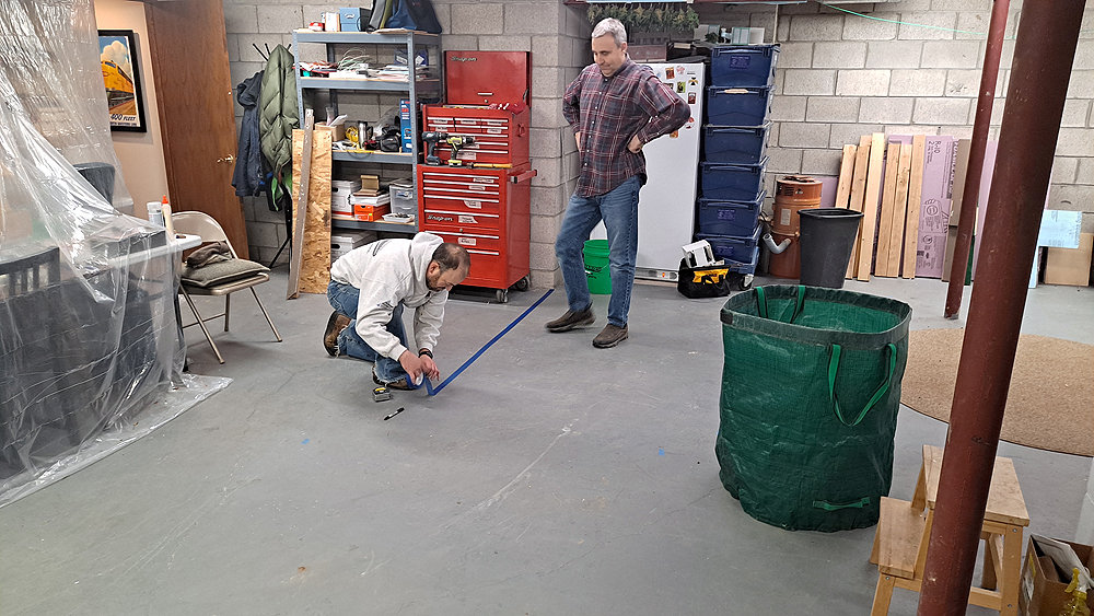 One man marks a location on a basement floor with blue masking tape while another one watches.