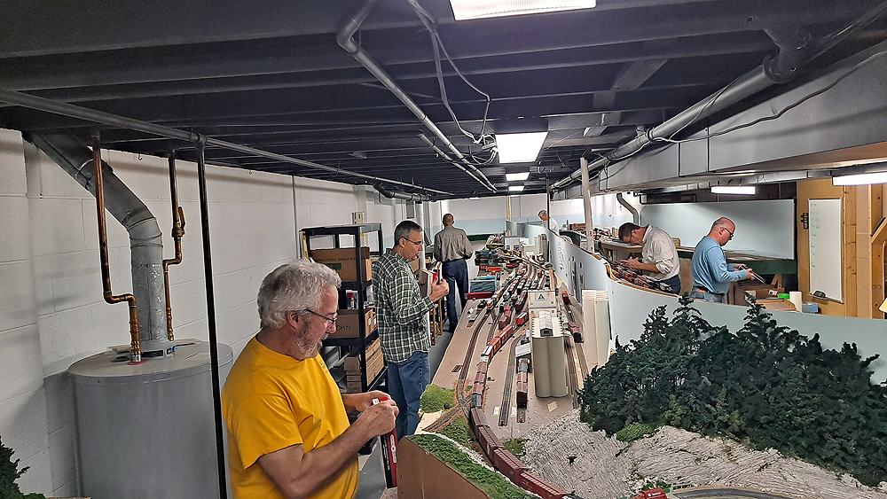 Packing up a model train layout: An island-style layout fills part of a basement with six people working around it.