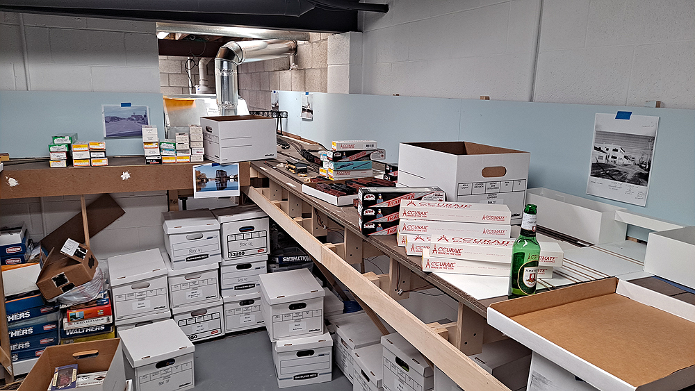 Rolling stock boxes are piled on the layout’s benchwork, and cardboard banker boxes are stacked below.