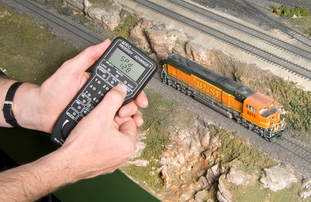 A hand holds a Digital Command Control throttle near a black-and-orange HO scale diesel on a scenicked layout