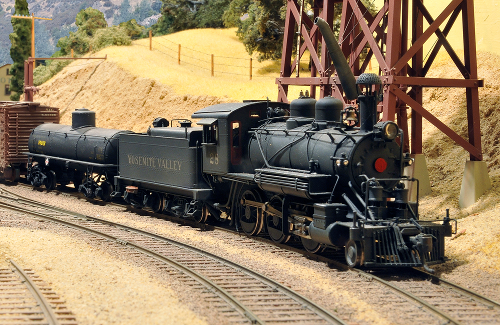 A short steam locomotive rolls around a curve alongside a water tank in a Western scene