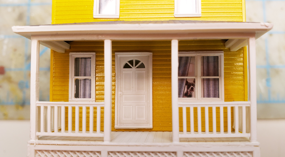 A yellow house with curtains, blinds, and dog in window