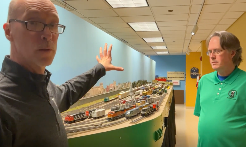 Two men facing a camera next to a model train layout.
