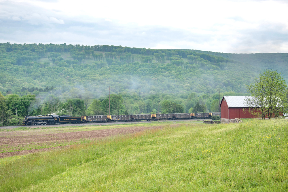 Steam locomotive and hopper cars seen from across field, passing barn