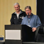 Man speaking at podium with smiling man standing to his right