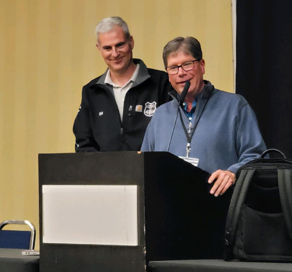 Man speaking at podium with smiling man standing to his right