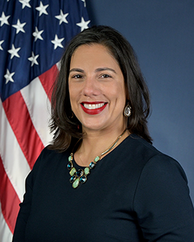 Woman with black hair in dark clothing standing in front of U.S. flag