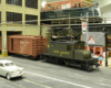 A green New Haven electric locomotive pushes a boxcar into a covered loading dock while in the background a larger one crosses a bridge.