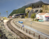 A blue-and-silver Amtrak train emerges from a tunnel and curves between a beach and a highway with a convenience store nearby