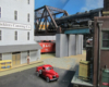 An articulated steam locomotive rumbles across a steel trestle above a street in an industrial area