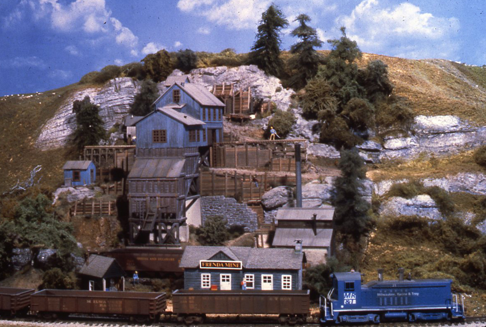  Photo of blue switcher and brown gondolas passing mine on hillside.