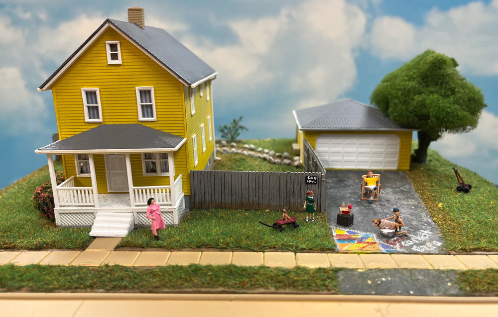 a diorama of a yellow and gray house with figures in the driveway
